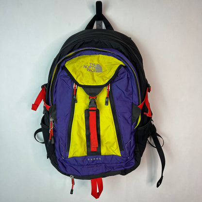 Vintage The North Face Surge 33L backpack in purple, yellow, and red colorblock design hanging on a white wall.