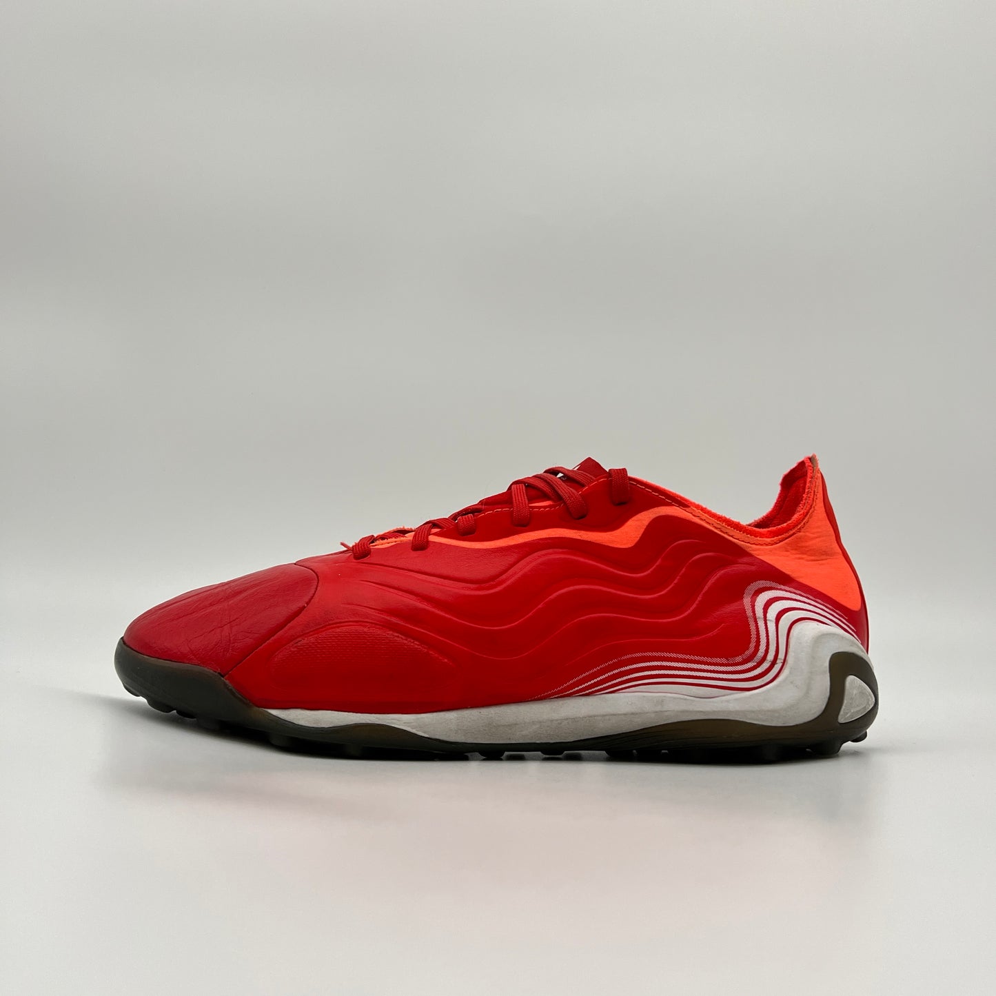 Side profile of a red adidas Copa Sense.1 turf shoe with wavy white heel details, red laces, and a dark grey studded outsole against a plain grey background.
