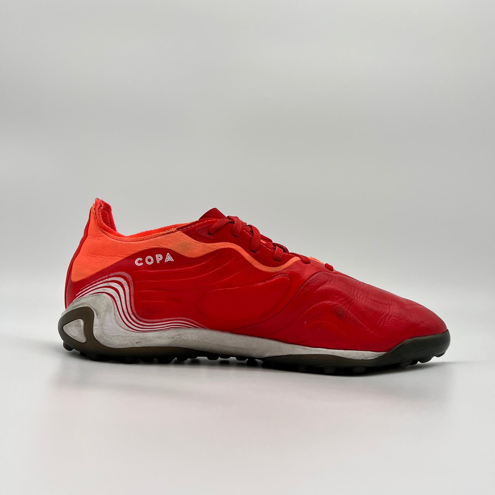 Side profile of a red adidas Copa Sense.1 turf shoe with wavy white heel details, red laces, and a dark grey studded outsole against a plain grey background.
