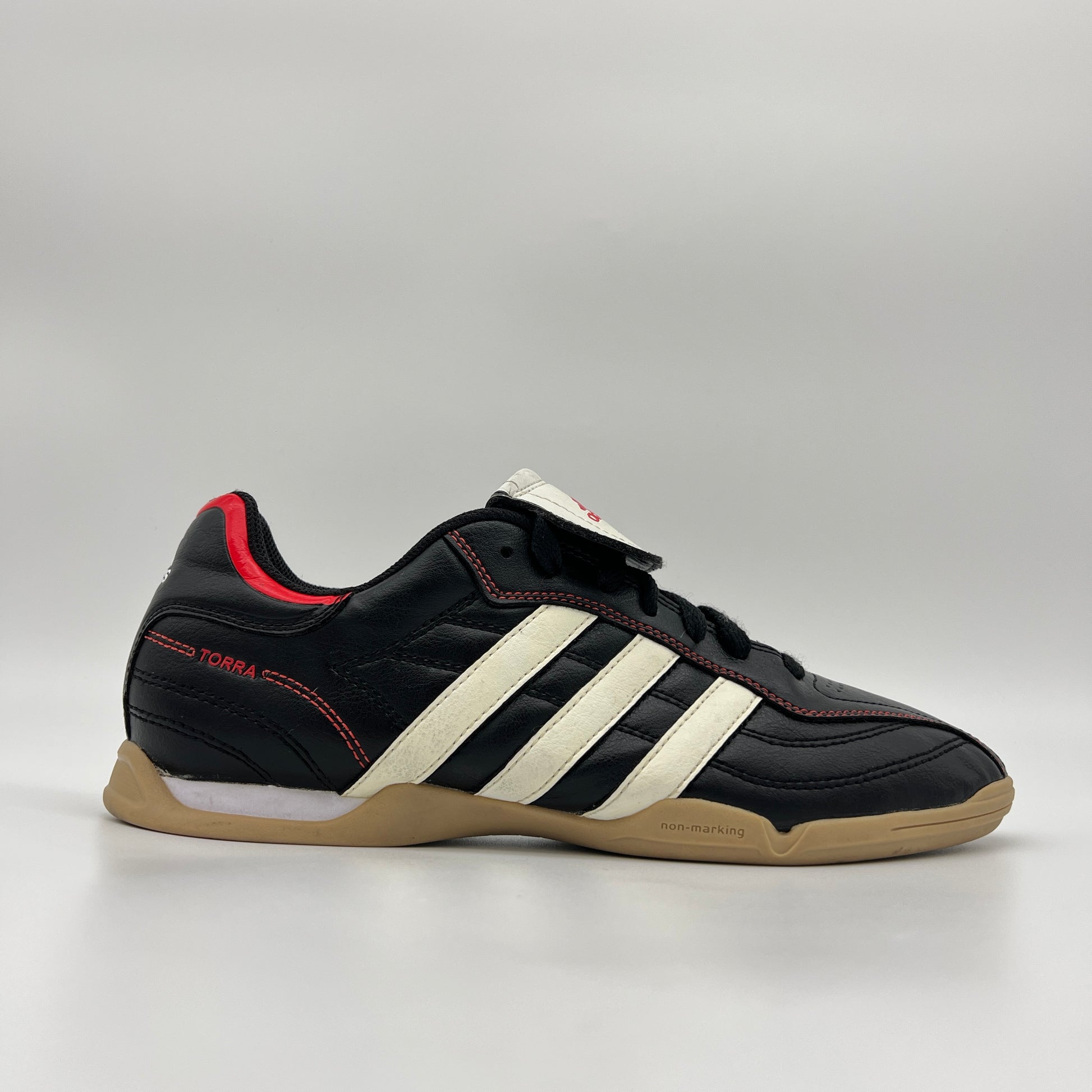 Side view of black Adidas Torra V IN J indoor soccer shoes with white stripes, red accents, and a gum rubber sole on a plain gray background.