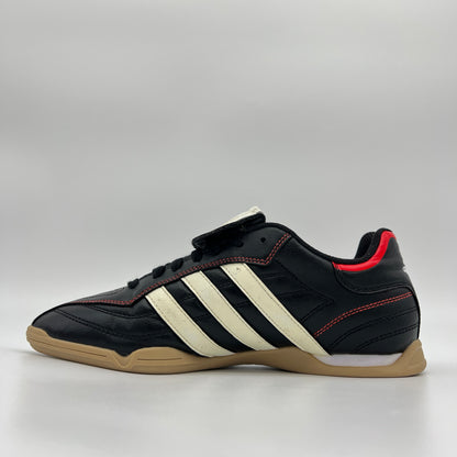 Side view of black Adidas Torra V IN J indoor soccer shoes with white stripes, red accents, and a gum rubber sole on a plain gray background.